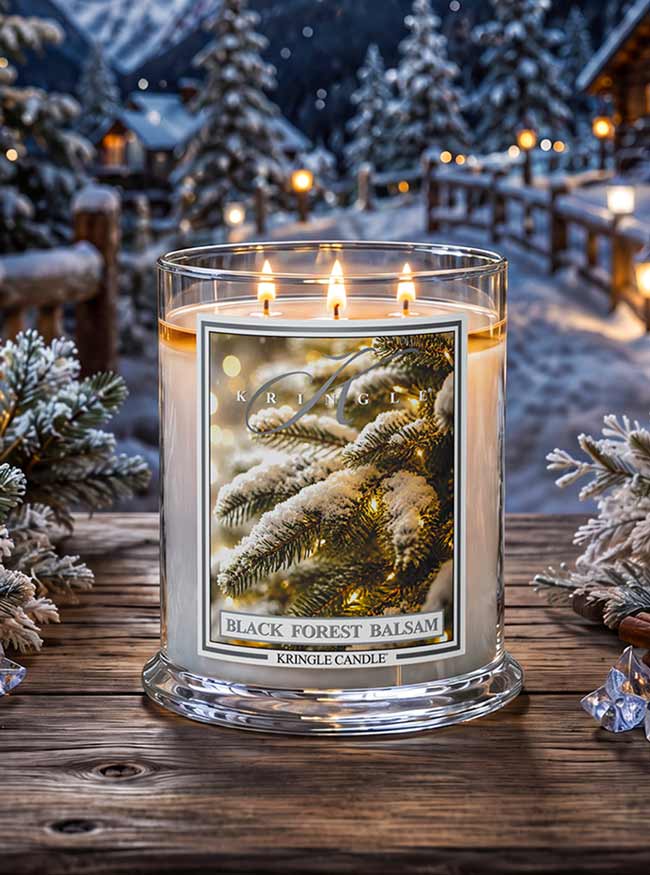 A three-wick glass jar candle labeled 'Black Forest Balsam' by Kringle Candle, lit and placed on a rustic wooden table with snowy pine trees and warm lights in the background.