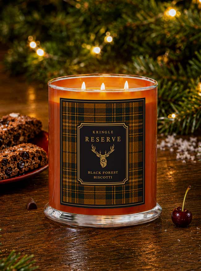A lit three-wick candle in a clear glass jar with amber-orange wax and a black-and-gold plaid label reading 'Kringle Reserve Black Forest Biscotti.' The candle sits on a wooden table with blurred Christmas greenery and warm lights in the background, plus cookies and a cherry nearby as styled props.