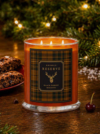 A lit three-wick candle in a clear glass jar with amber-orange wax and a black-and-gold plaid label reading 'Kringle Reserve Black Forest Biscotti.' The candle sits on a wooden table with blurred Christmas greenery and warm lights in the background, plus cookies and a cherry nearby as styled props.