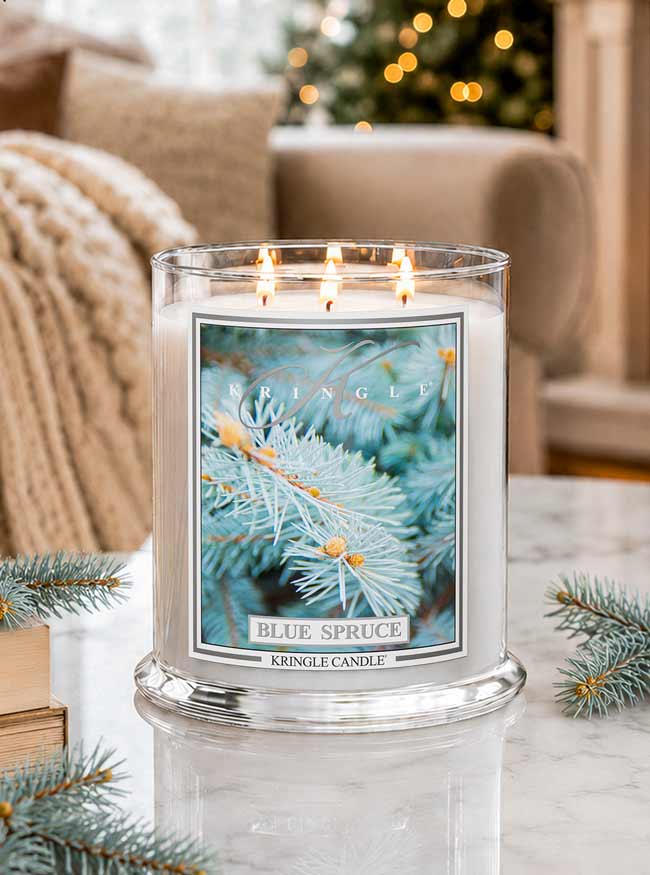 A lit Kringle Candle in a clear glass jar with three flames, featuring a blue spruce pine branch label, set on a marble table with holiday greenery and a cozy living room background.