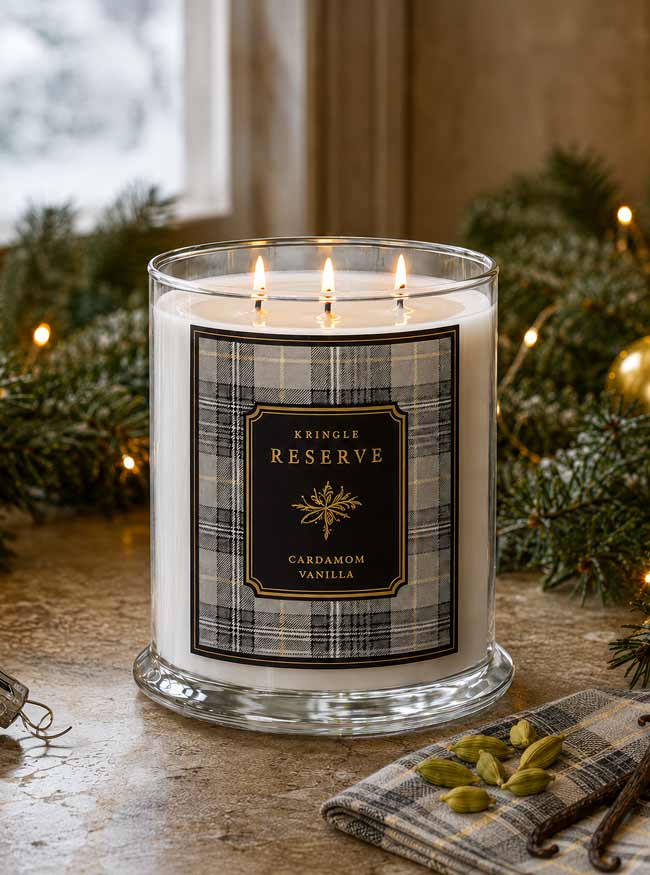 A three-wick candle in a clear glass jar sits on a stone surface with lit flames. The candle has a cream-colored wax and a front label reading 'Kringel Reserve Cardamom Vanilla' in a plaid pattern.