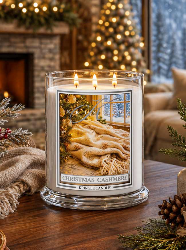 A lit three-wick candle in a clear glass jar sits on a wooden table in a cozy holiday living room. The candle label reads 'CHRISTMAS CASHMERE' and shows a warm image of a knitted beige throw blanket in front of a window.