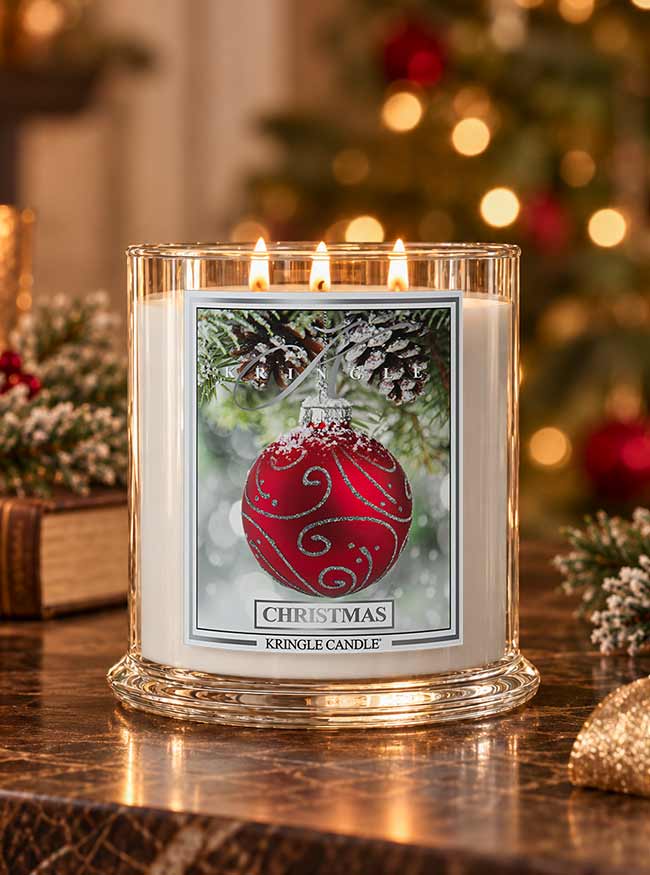 A lit Kringle Candle in a clear glass jar sits on a reflective wooden table with a blurred Christmas tree and holiday decorations in the background. The candle features a front label with an illustration of a red ornament on a snowy branch and the text 'CHRISTMAS' and 'KRINGLE CANDLE'.