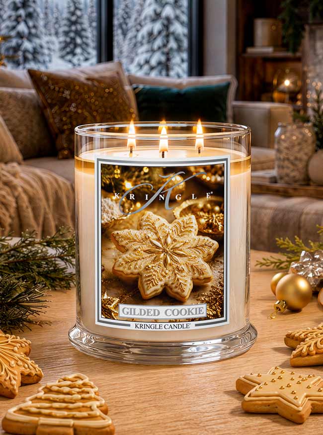 A Kringle Candle in a clear glass jar with three lit wicks, labeled 'Gilded Cookie,' placed on a wooden table in a cozy living room setting with holiday decorations and cookies.