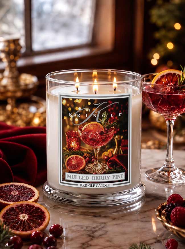 A lit Kringle Candle in a clear glass jar on a marble surface, surrounded by cranberries, orange slices, and a red cocktail glass in a warm holiday setting. The front label reads 'Mulled Berry Pine' and shows a festive illustration.