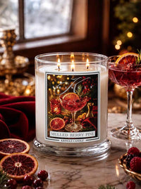 A lit Kringle Candle in a clear glass jar on a marble surface, surrounded by cranberries, orange slices, and a red cocktail glass in a warm holiday setting. The front label reads 'Mulled Berry Pine' and shows a festive illustration.
