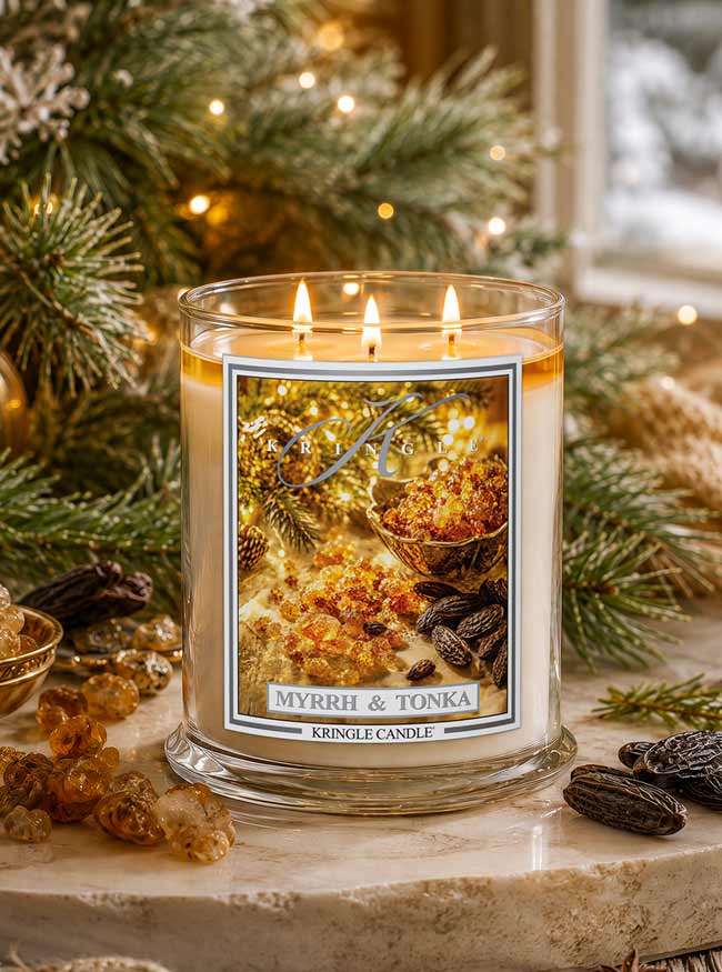 A lit double-wick candle in a clear glass jar sits on a stone surface surrounded by pine branches, amber crystals, and dried botanicals. The front label reads 'MYRRH & TONKA' and 'KRINGLE CANDLE'.