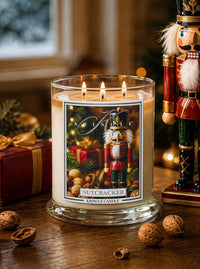A lit Kringle Candle in a clear glass jar with three flames, featuring a festive nutcracker illustration on the label. The candle sits on a wooden table surrounded by holiday decor, including a wrapped gift, scattered walnuts, and a large nutcracker figurine in the background.