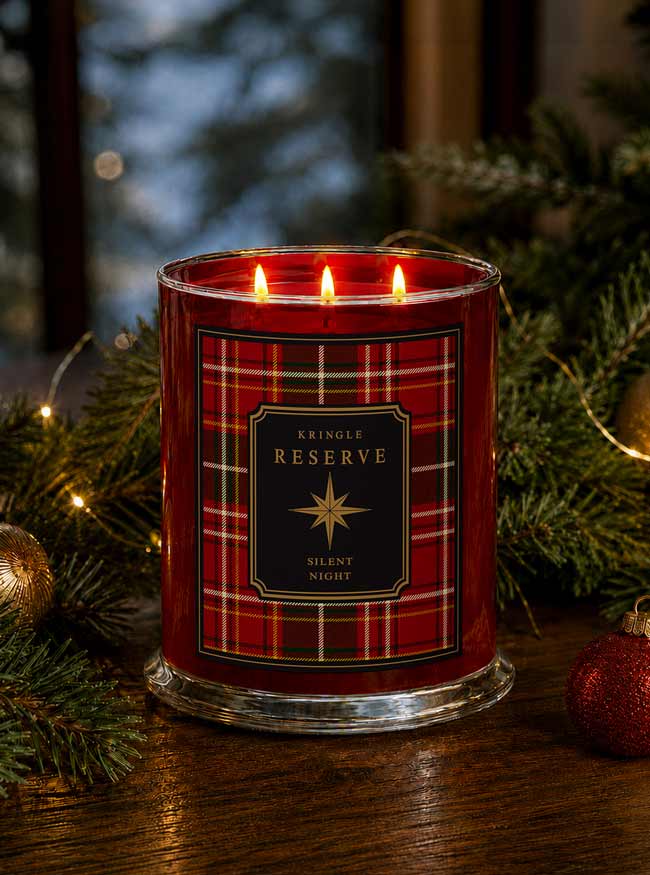 A red holiday-themed candle in a clear glass jar with three lit wicks, featuring a red plaid label that reads 'Kringle Reserve Silent Night.' The candle is styled on a wooden surface with evergreen branches, warm string lights, and red ornaments in the background.