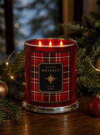 A red holiday-themed candle in a clear glass jar with three lit wicks, featuring a red plaid label that reads 'Kringle Reserve Silent Night.' The candle is styled on a wooden surface with evergreen branches, warm string lights, and red ornaments in the background.