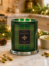 A three-wick scented candle in a clear glass jar with a green plaid label reading 'Kringle Reserve Velvet Pistachio Creme,' lit on a marble surface with holiday greenery, nuts, and a bowl nearby.