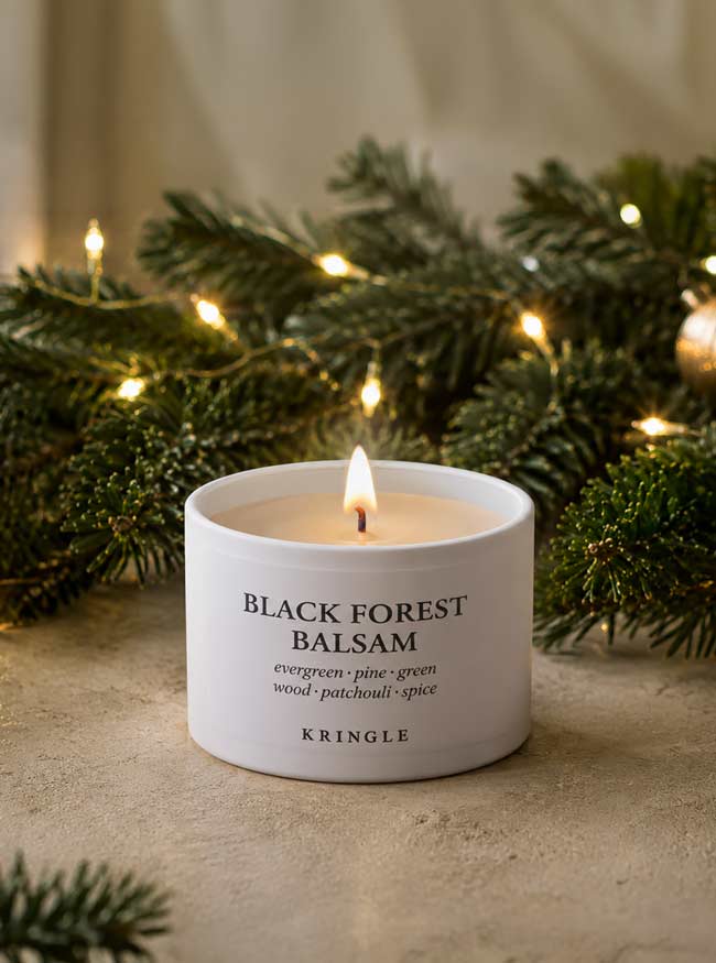 A lit white ceramic candle labeled 'BLACK FOREST BALSAM' by Kringle, set on a stone-like surface with pine branches and warm string lights in the background.