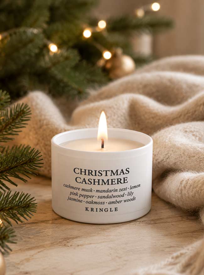 A lit white candle labeled 'CHRISTMAS CASHMERE' by Kringle sits on a marble surface with a cozy beige knit blanket and blurred Christmas tree lights in the background.