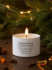 A lit white candle in a cylindrical jar labeled 'CINNAMON CLOVE ZEST' by Kringle, placed on a dark wooden surface with cinnamon sticks, orange peel, and evergreen branches in the background.
