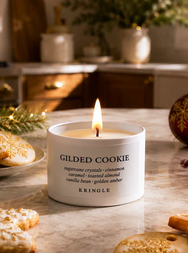 A lit white candle labeled 'GILDED COOKIE' by Kringle sits on a marble countertop with holiday cookies, evergreen branches, and warm blurred background decor.