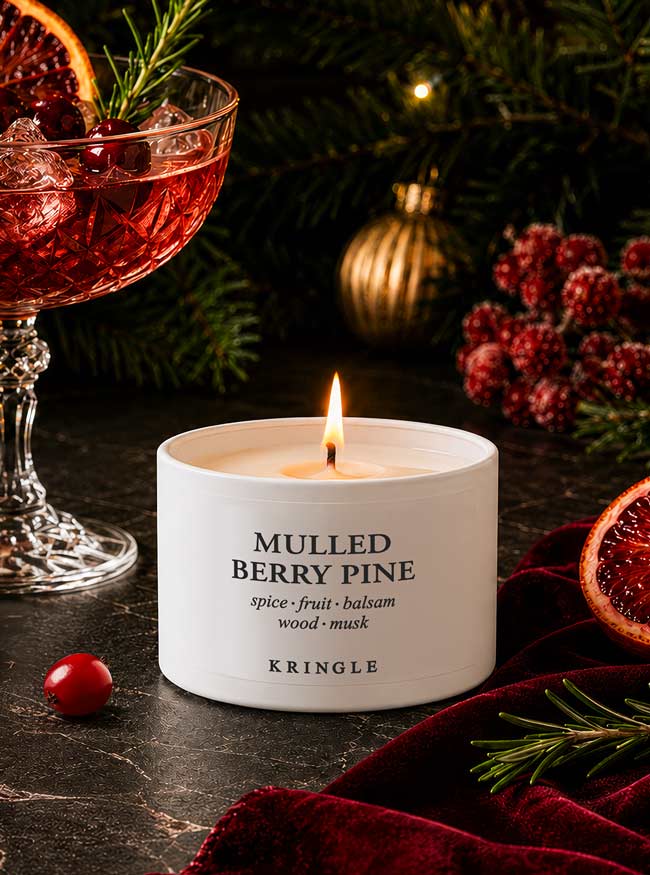 A white Kringle candle labeled 'Mulled Berry Pine' is lit on a dark tabletop with holiday decor, including pine branches, cranberries, a glass of mulled wine, and a red velvet cloth.