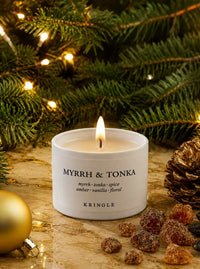 A lit white candle labeled 'MYRRH & TONKA' by Kringle, styled on a stone surface with pine branches, warm string lights, a gold ornament, and dried fruit in the background.