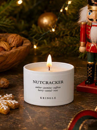 A white ceramic Kringle Nutcracker candle with a lit flame, set on a rustic wooden table with a nutcracker, walnuts, and holiday lights in the background.