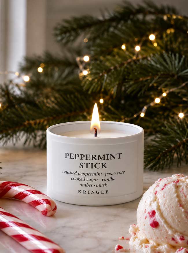 A lit white candle labeled 'PEPPERMINT STICK' by KRINGLE sits on a marble surface with candy canes and a peppermint ice cream scoop in the foreground, and blurred evergreen branches with warm string lights in the background.