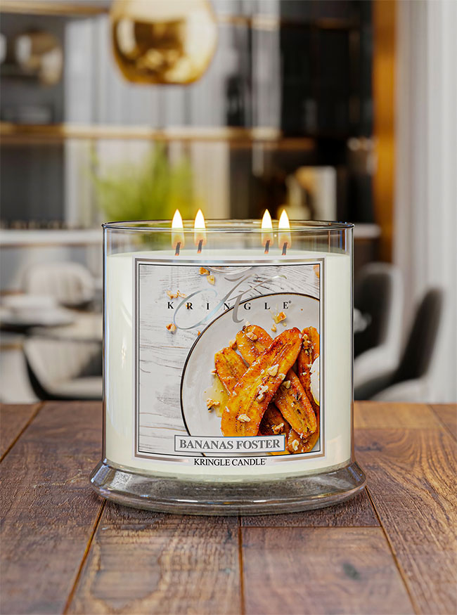 A lit Kringle Candle in a clear glass jar with three flames, featuring a 'Bananas Foster' scent label with a photo of banana slices on a plate, set on a wooden table with a blurred kitchen background.