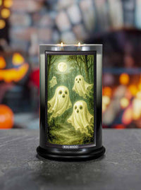 Halloween-themed Boo-Whoo candle with ghost design on a dark surface, blurred pumpkins and jack-o'-lanterns in the background.