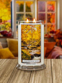 Kringle Candle with 'Amberwood Moss' candle on a wooden surface with a blurred autumn-themed background.