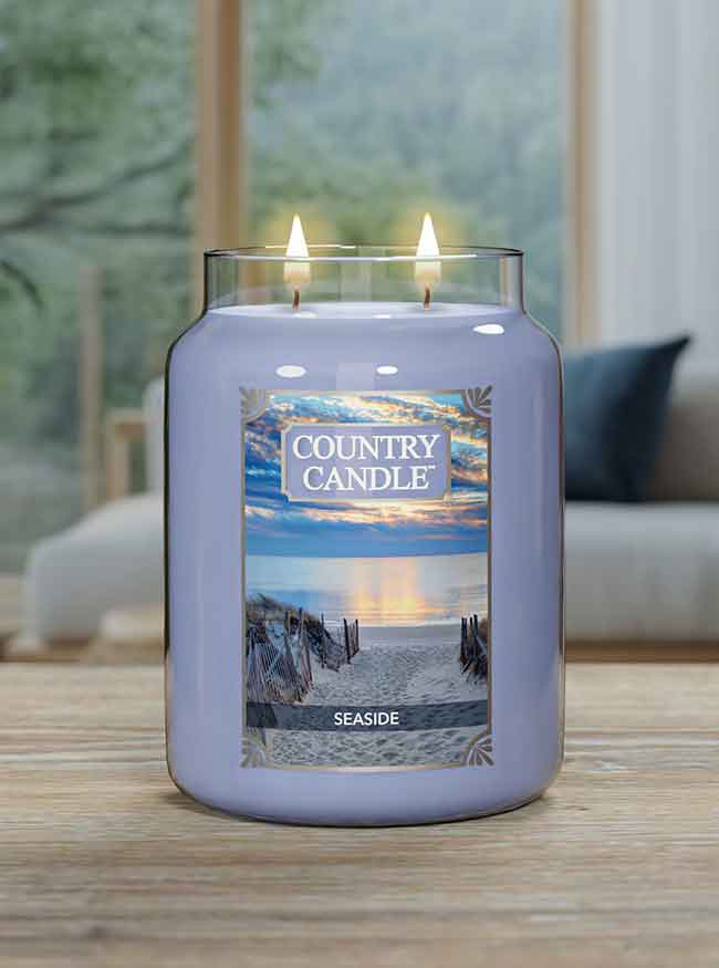 Lit two-wick candle labeled "Country Candle Seaside" on a wooden table with a blurred living room background.
