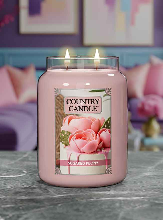 A lit jar candle labeled "Country Candle, Sugared Peony" on a marble surface with a cozy, colorful background.