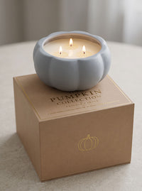 A three-wick scented candle in a ribbed light blue-gray pumpkin-shaped holder sits on top of a kraft paper box labeled 'Pumpkin Collection' with a gold pumpkin outline, set on a neutral tabletop.
