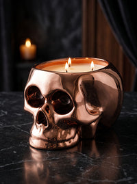 A metallic copper skull-shaped candle holder with three lit wicks, set on a dark marble surface with a blurred dark background.