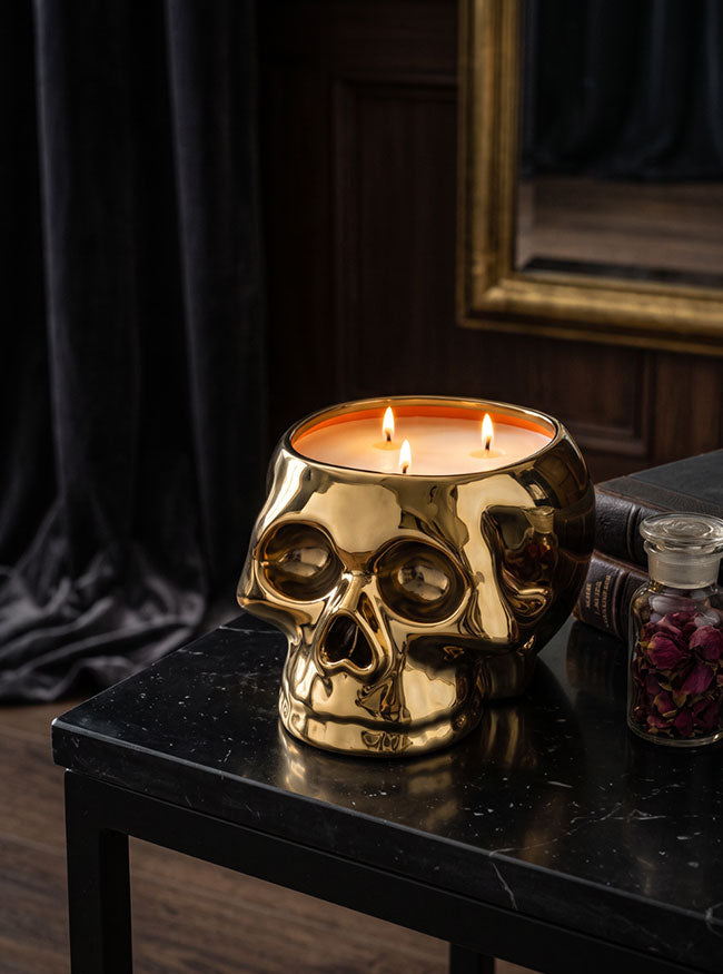 A gold skull-shaped candle holder with three lit candles, placed on a black marble table in a dark, moody room with a mirror and black curtain in the background.