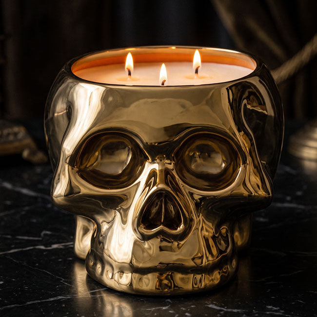 A metallic gold skull-shaped candle holder with three lit wicks, set on a dark marble surface against a dark, moody background.