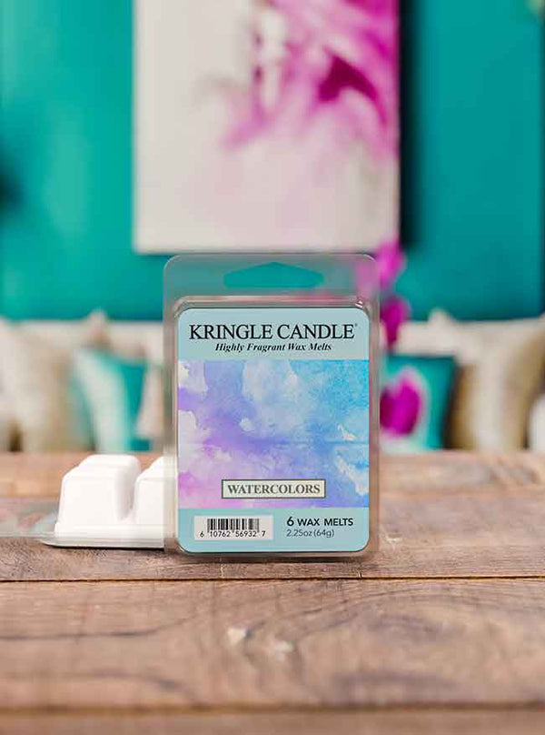 A package of Kringle Candle “Watercolors” highly fragrant wax melts displayed on a rustic wooden surface. The label features a soft watercolor design in pastel shades of purple, blue, and pink. An open wax melt tray with white cubes sits beside the package. The background includes a teal wall, floral artwork, and decorative pillows, creating a stylish and vibrant room setting.