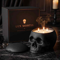 A black skull-shaped candle with three lit wicks sits on a dark marble surface beside its branded box. The box reads 'LUX MORTIS BY KRINGLE CANDLE' with a small skull logo.
