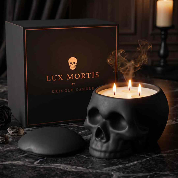 A black skull-shaped candle with three lit wicks sits on a dark marble surface beside its branded box. The box reads 'LUX MORTIS BY KRINGLE CANDLE' with a small skull logo.