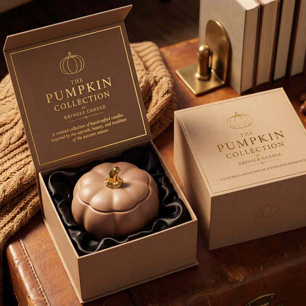 An open and closed kraft paper box for The Pumpkin Collection single candle, showing a pumpkin-shaped candle in black satin packaging. The scene is styled on a rustic leather surface with a knitted sweater and books in the background.