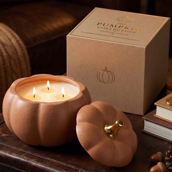 A pumpkin-shaped candle in a terracotta-colored pot with three lit wicks, shown beside its matching lid and branded box on a dark wooden surface. The box reads 'The Pumpkin Collection Scented Candle' and features a gold pumpkin outline.