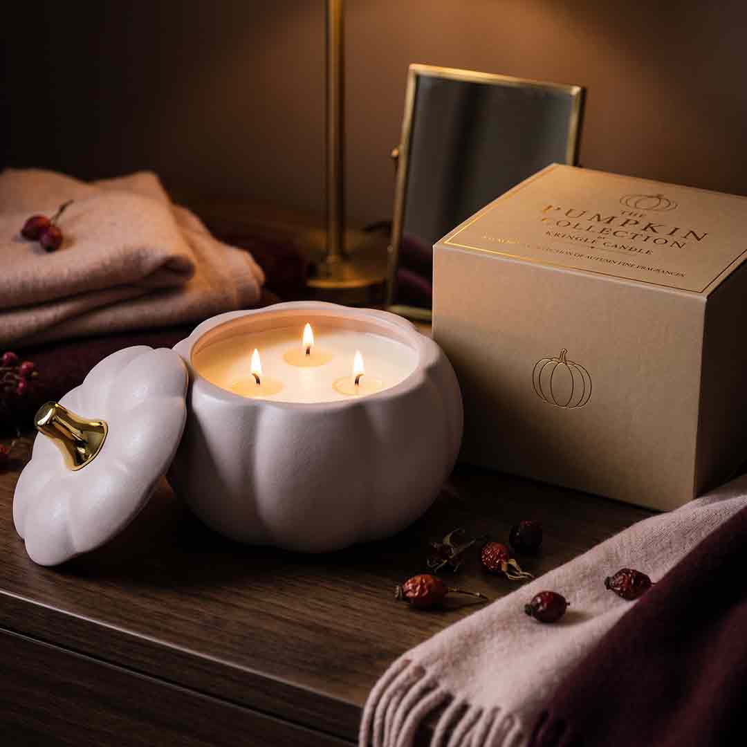A pumpkin-shaped candle jar with its lid off, showing three lit wicks, placed on a wooden table beside its branded box. The scene includes folded blankets, dried berries, and a mirror in a warm, cozy indoor setting.