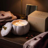 A pumpkin-shaped candle jar with its lid off, showing three lit wicks, placed on a wooden table beside its branded box. The scene includes folded blankets, dried berries, and a mirror in a warm, cozy indoor setting.