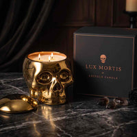 A gold skull-shaped candle with three lit wicks sits on a dark marble surface beside its black branded box. The box reads 'LUX MORTIS by Kringle Candle' with a small skull logo.