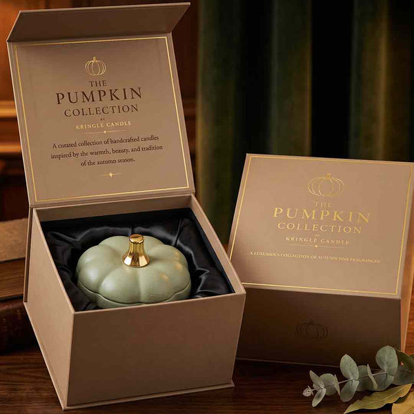 A pumpkin-shaped candle in a pale green ceramic pot with a gold top, presented in an open beige gift box labeled 'The Pumpkin Collection Kringle Candle.' A matching closed box sits beside it on a wooden surface with a blurred indoor background.