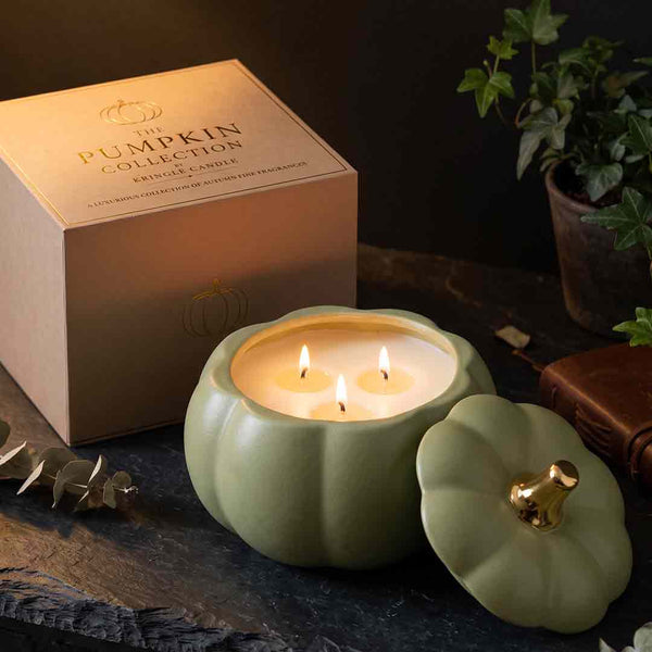 A pumpkin-shaped candle in a matte sage green container with its lid placed beside it, showing three lit wicks and warm candlelight. The branded box reading 'Pumpkin Collection' sits behind it on a dark tabletop, with a potted plant and leather item in the background.