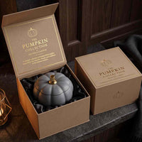 A gray pumpkin-shaped candle presented in an open kraft gift box with gold lettering, alongside a closed matching box on a dark wooden surface.
