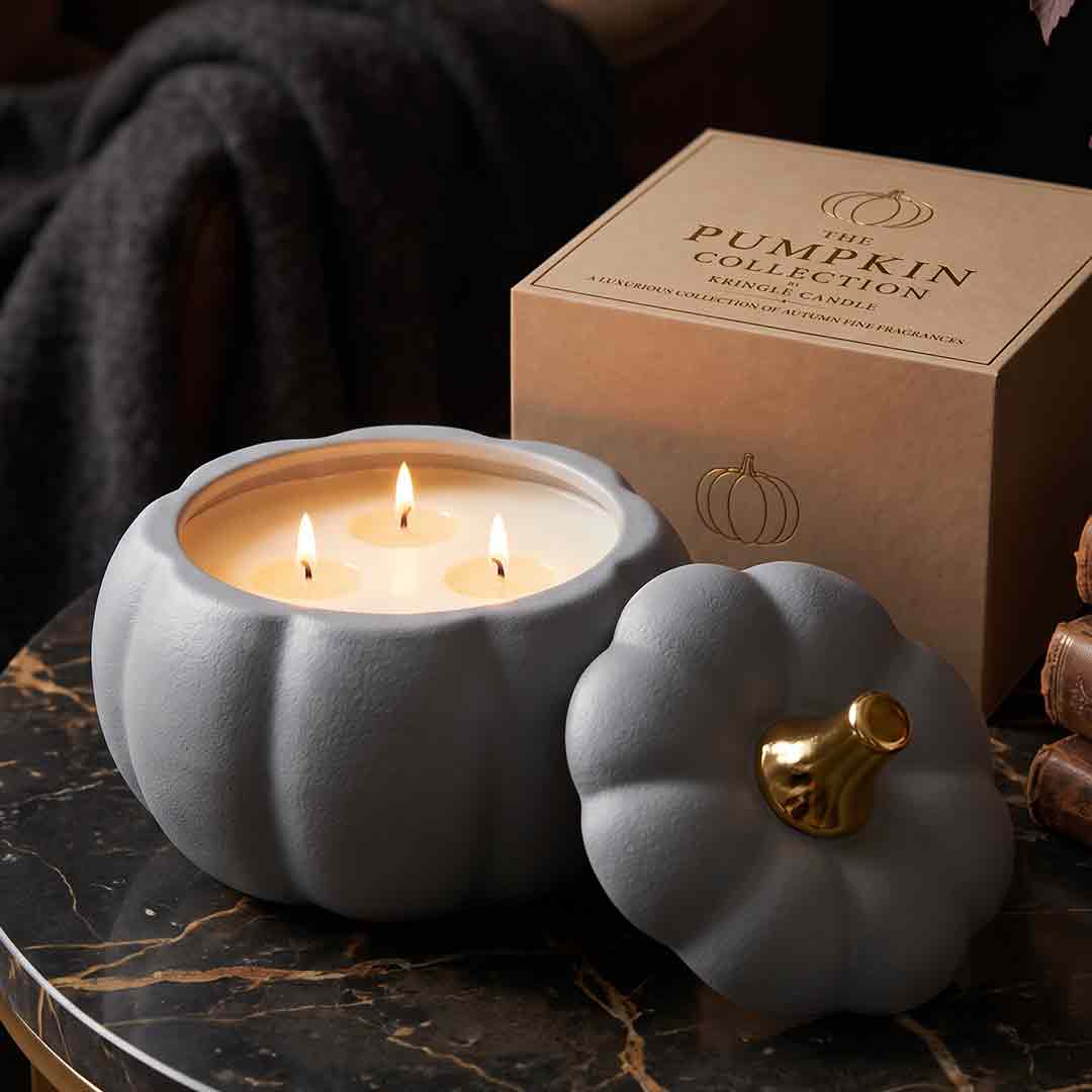 A pumpkin-shaped candle holder with three lit candles in front of its kraft paper box, styled on a dark marble surface with a cozy, dark background.