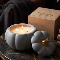 A pumpkin-shaped candle holder with three lit candles in front of its kraft paper box, styled on a dark marble surface with a cozy, dark background.