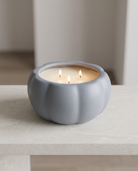 A lit two-wick candle in a matte gray pumpkin-shaped holder, resting on a light stone surface with a softly blurred indoor background.
