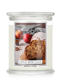 A Kringle Candle jar candle with a clear glass lid and base. The front label features a photo of sliced apple rum cake with apples and spices, and the text 'KRINGLE' and 'APPLE RUM CAKE' plus 'KRINGLE CANDLE'.