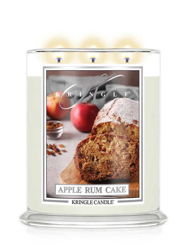 A Kringle Candle in a clear glass jar with three lit wicks, featuring a front label that reads 'Apple Rum Cake' and shows a loaf and sliced cake with apples on a light background.