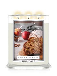 A Kringle Candle in a clear glass jar with three lit wicks, featuring a front label that reads 'Apple Rum Cake' and shows a loaf and sliced cake with apples on a light background.