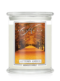 A Kringle Candle in a clear glass jar with cream-colored wax and a front label depicting an autumn tree-lined path. The label reads 'KRINGLE', 'AUTUMN AMBER', and 'KRINGLE CANDLE'.